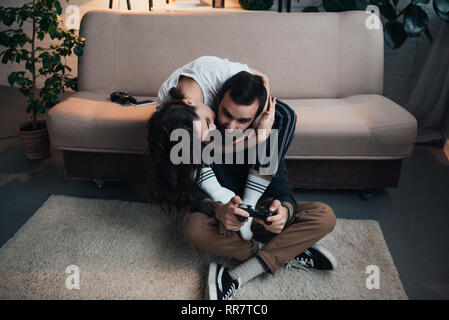 woman hugging and kissing man playing video game with joystick in living room Stock Photo