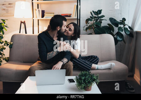 beautiful young couple sitting on couch and kissing near laptop and smartphone on table in living room Stock Photo