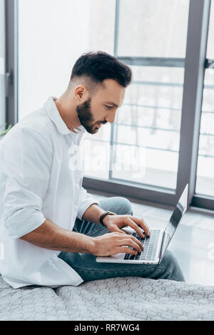 Concentrated handsome man typing on laptop keyboard in bedroom Stock Photo