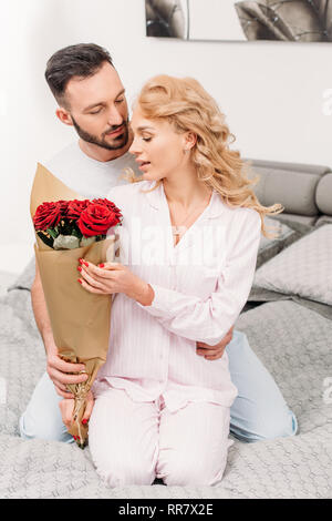 Man presenting flowers to girlfriend while sitting on bed Stock Photo