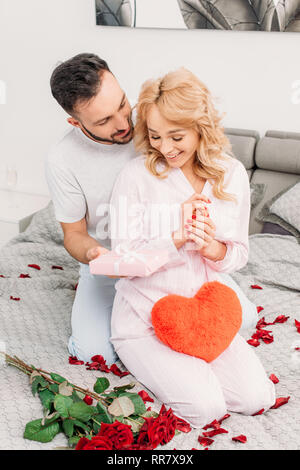 Smiling man sitting on bed with roses and presenting gift to girlfriend Stock Photo