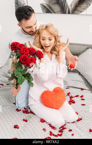 Brunette man presenting flowers and ring box to surprised girlfriend in bedroom Stock Photo