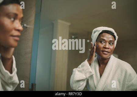 Reflection of mature african woman in bathroom mirror applying cosmetic cream on her face. Mature female in bathrobe putting on cream on her face skin Stock Photo