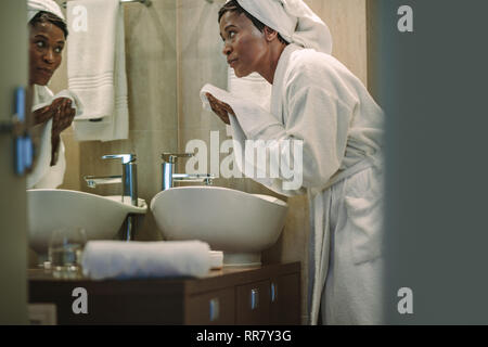 African female cleaning her face with towel in bathroom. Mature woman wearing bathrobe standing in front of mirror and wiping her face with towel. Stock Photo