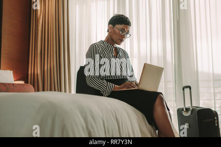 Businesswoman sitting on bed using laptop. African woman on business trip working in hotel room. Stock Photo