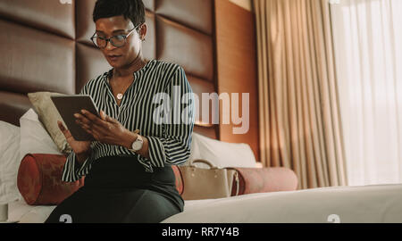 Businesswoman sitting on bed using digital tablet. African woman on business trip working in hotel room. Stock Photo
