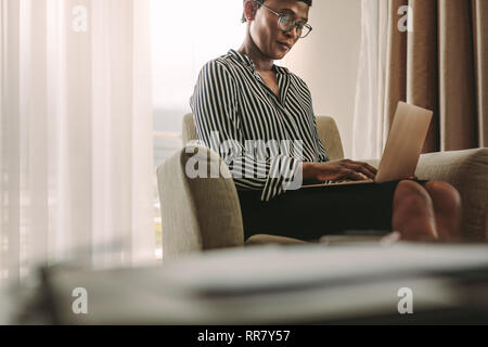 African woman in formal clothes sitting on armchair in hotel room using laptop. Businesswoman on business trip working from hotel room. Stock Photo
