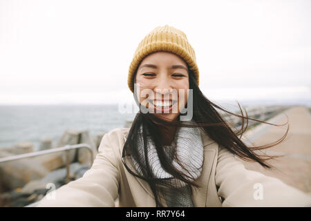 Close up of a smiling woman in winter wear standing near the sea. Asian woman standing outdoors on a cold winter morning with open arms. Stock Photo