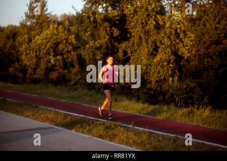 one young woman, 20-29 years, running alone outdoors in Summer, on a red tartan running track. Warm dawn, sunset light. Stock Photo