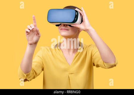 Young woman in her 30s using virtual reality goggles. Woman wearing VR glasses isolated over yellow background. VR experience concept. Stock Photo