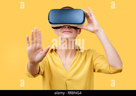Young woman in her 30s using virtual reality goggles. Woman wearing VR glasses isolated over yellow background. VR experience concept. Stock Photo