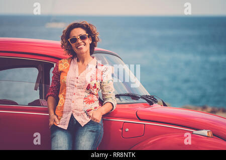 Beautiful caucasian middle age lady driver pose standing with a red legendary retro vintage car behind - ocean in background for travel and alternativ Stock Photo