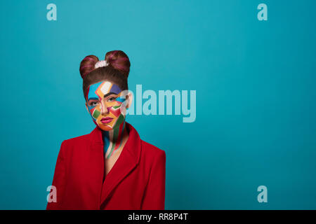 Stylish, trendy model wearing in red jacket, posing in studio with blue background. Cool girl has creative pop art make up on face and nice bow hairst Stock Photo