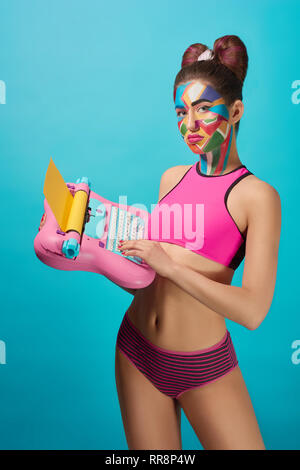 Stylish girl with slender figure looking at camera, holding pink toy typewriter, standing. Beautiful, cool model has pop art colourful make up on face Stock Photo