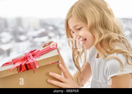 Beautiful girl with long, blonde, curly hair smiling and opening big craft box with red bow. Happy daughter receiving present on holiday and rejoicing Stock Photo
