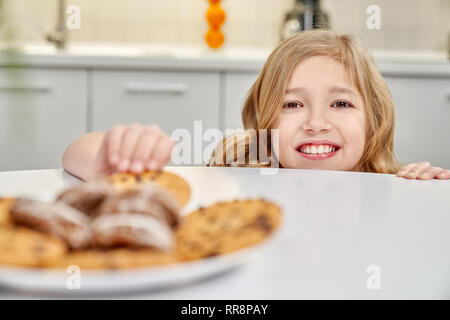 Delicious american cookies on big white plate. Tasty biscuits with raisins and chocolate cookies on white table. Child hiding behind table and secretl Stock Photo