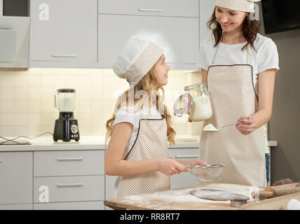 Young mother and pretty daughter in white and brown aprons, chef hats. Girl with long hair and woman sifting flour, holding spoon and flour jar and co