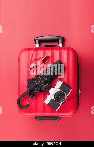 Red travel suitcase with red sunglasses, old camera, black umbrella on red background. Minimal style Stock Photo