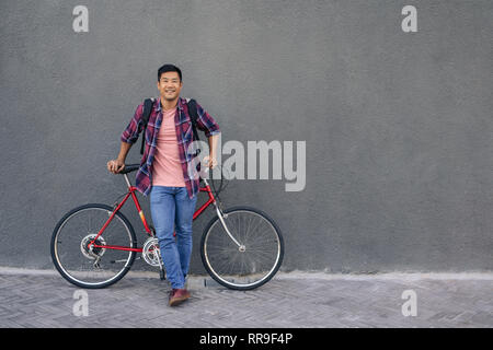 Smiling young man standing with his bike against a wall Stock Photo