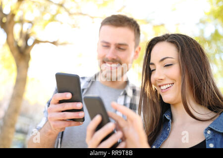 Two happy friends using their smart phones standing outdoors in a park Stock Photo