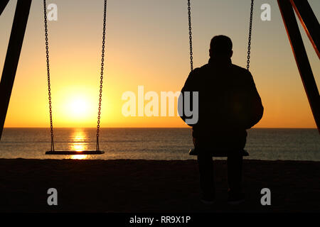 Back view backlighting silhouette of a man sitting on swing alone looking at sunset on the beach