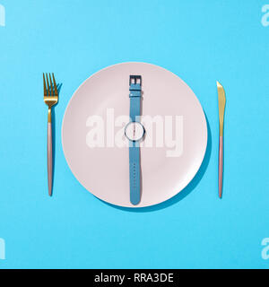 White plate with whatch shows six o'clock served knife and fork on a blue background and shadows. Time to eat and diet concept. Top view. Stock Photo