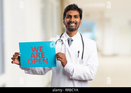 Male indian physician or doctor holding we are open sign text on blue paper with friendly expression Stock Photo