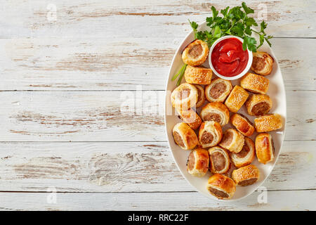 Delicious homemade sausage rolls on a white oval platter with tomato sauce on a wooden rustic table, finger food, english cuisine, view from above, fl Stock Photo