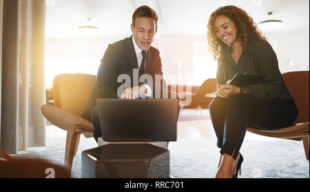 Two diverse young businesspeople discussing work and using a laptop while sitting together in a modern office Stock Photo