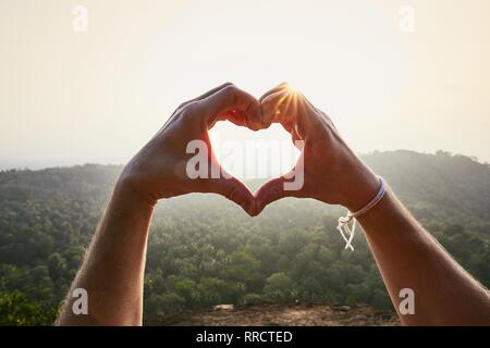 Close-Up of hands making heart shape against landscape at sunset. Stock Photo
