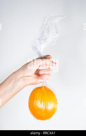Excessive plastic use concept: orange in plastic wrap held in a hand. Unreasonably over-packaged food products: person holds fresh fruit in kitchen wr Stock Photo