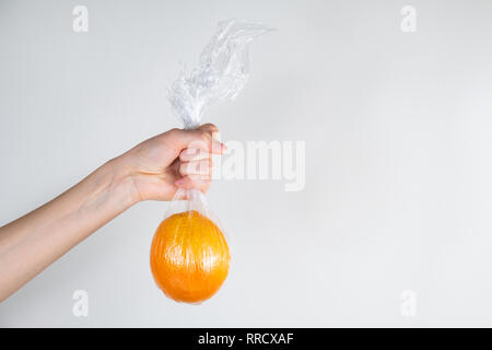 Excessive plastic use concept: orange in plastic wrap held in a hand. Unreasonably over-packaged food products: person holds fresh fruit in kitchen wr Stock Photo