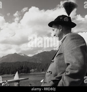 1950s, German man  wearing a traditional Bavarian jacket and Tyrolean hat, with feather, looking over an alpine lake, Germany. Stock Photo