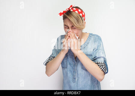Portrait of sick young woman in casual blue denim shirt with red headband standing with tissue and cleaning her runny nose. indoor studio shot, isolat Stock Photo