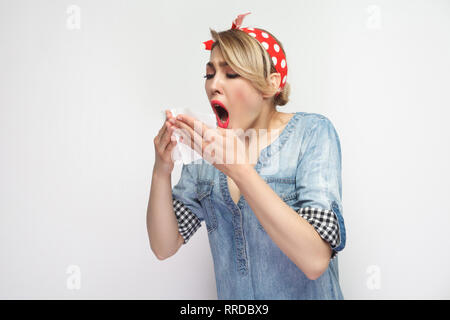 Portrait of sick young woman in casual blue denim shirt with makeup and red headband standing, holding tissue and sneezing. indoor studio shot, isolat Stock Photo