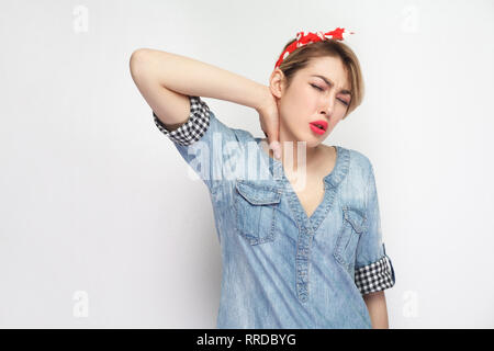 Neck pain. Portrait of sad young woman in casual blue denim shirt with makeup and red headband standing, closed eyes and holding her painful neck. ind Stock Photo