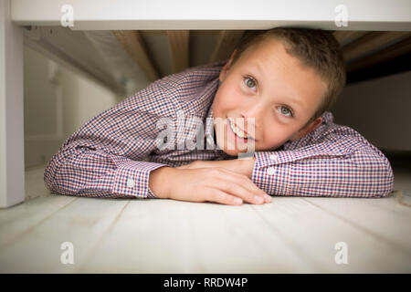 Portrait of a smiling boy lying on his stomach while hiding underneath a wooden bed. Stock Photo
