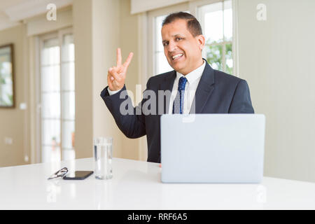 Middle age business man working with computer laptop smiling with happy face winking at the camera doing victory sign. Number two. Stock Photo
