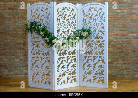 Folding wooden screen or room divider with three panel partition, embellished with a flower garland and set against a brick wall Stock Photo