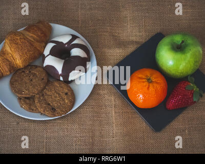 A plate of black slate with a green apple, a tangerine and a strawberry and one plate with one croissant, one chocolate donut and cookies on a burlap  Stock Photo