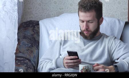 One white young brunette curly-bearded man uses the phone sitting in a chair drinking tea or coffee Stock Photo