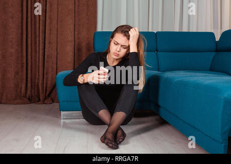 Jealous woman sitting near sofa holding phone feeling sad waiting for call, frustrated millennial girl upset, worried receiving bad news in mobile mes Stock Photo