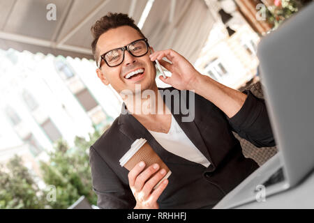 Lunch time. Young man in suit and glasses sitting at cafe on city street with laptop with cup of coffee talking on smartphone laughing cheerful Stock Photo