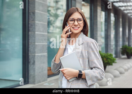 Business communications. Young lady in eyeglasses standing on the city street with digital tablet talking on smartphone cheerful Stock Photo
