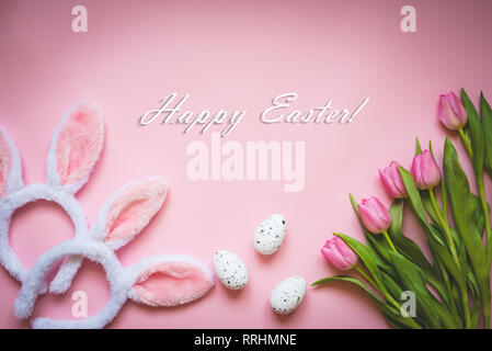 Top view of Easter eggs, pink tulips and two white fluffy bunny ears over pink background. Easter concept background. Happy Easter text. Stock Photo