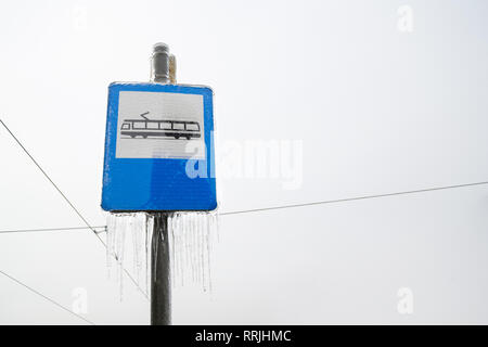 Tram station sign with icicles hanging from it, on blue background, during Winter, with an overcast sky in the back and electricity wires spread acros Stock Photo
