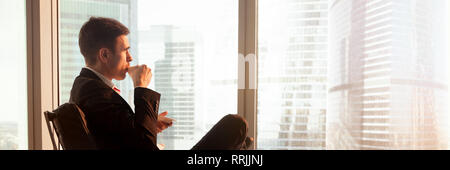 Horizontal view relaxed businessman sitting drinking coffee looking at window Stock Photo