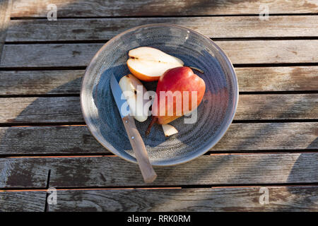 Red pears on blue plate cut in slices by knife in sunlight on wooden table. Stock Photo