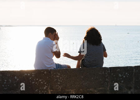 Rear view of couple fooling around while sitting on wall during sunset against ocean Stock Photo
