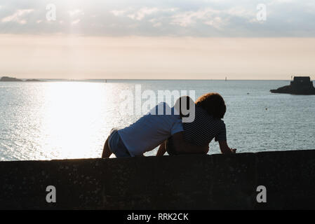 Rear view of couple fooling around while sitting on wall during sunset against ocean Stock Photo